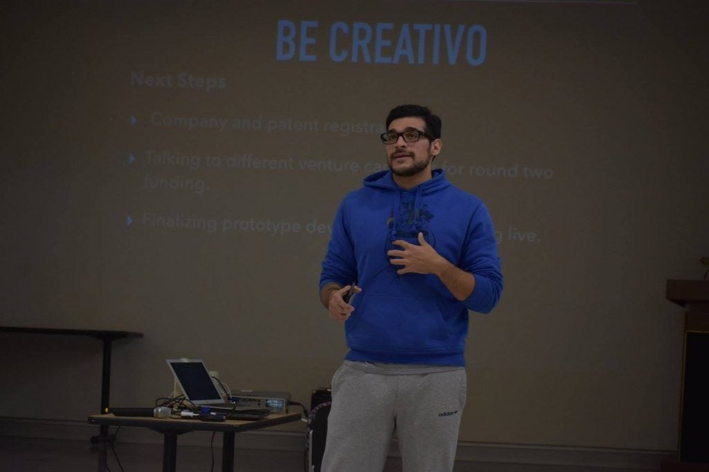 Student in blue sweatshirt talking in front of a screen with the words "Be Creativo" on it.