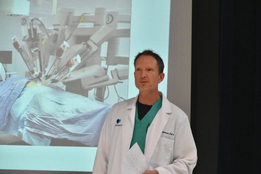 Man in lab coat speaking while a picture of a surgery is on a screen.